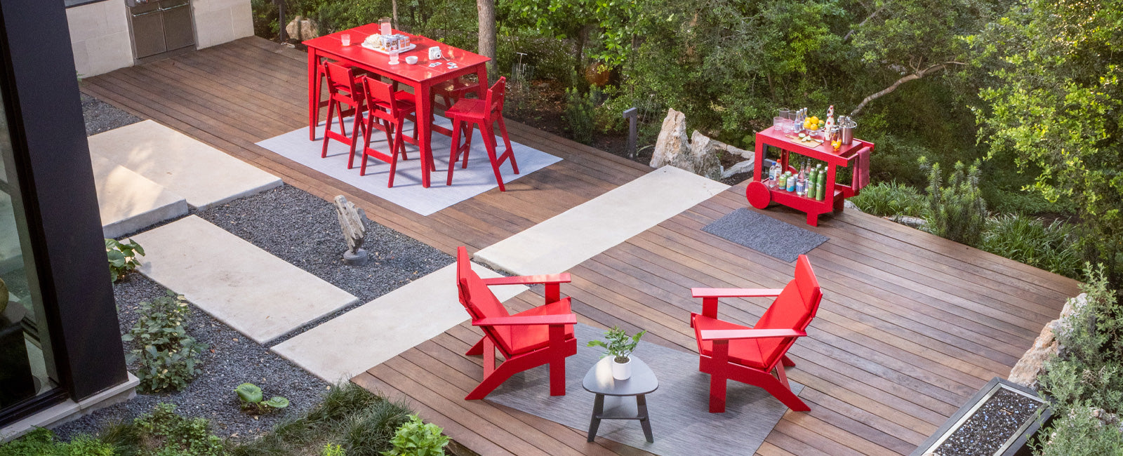 overhead image of wooden patio. A red alfresco bar table with bar stools is in upper left corner. A red lollygagger bar cart is to the right of the bar table along patio edge. bottom is a pair of red lollygagger lounge chairs with black rapson side table.