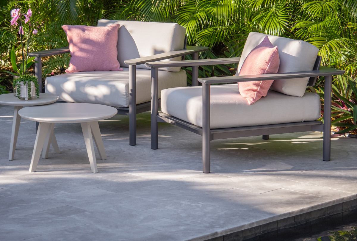 two charcoal aurora lounge chairs with cloud cushions near a tropical poolside. A satellite side table and end table are placed in front of the chairs. 